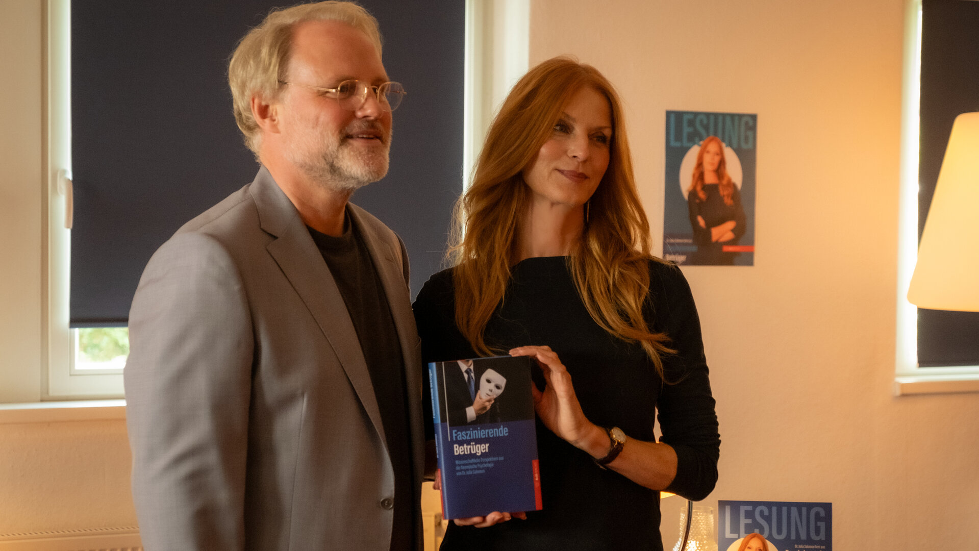 "Blutige Anfänger - Edelfeder": Henry Brill (Mirco Reseg) und Dr. Julia Salomon (Esther Schweins) stehen dicht nebeneinander in einem Raum. Julia hält ihr Buch vor ihrem Körper. Beide blicken lächelnd zur Seite.