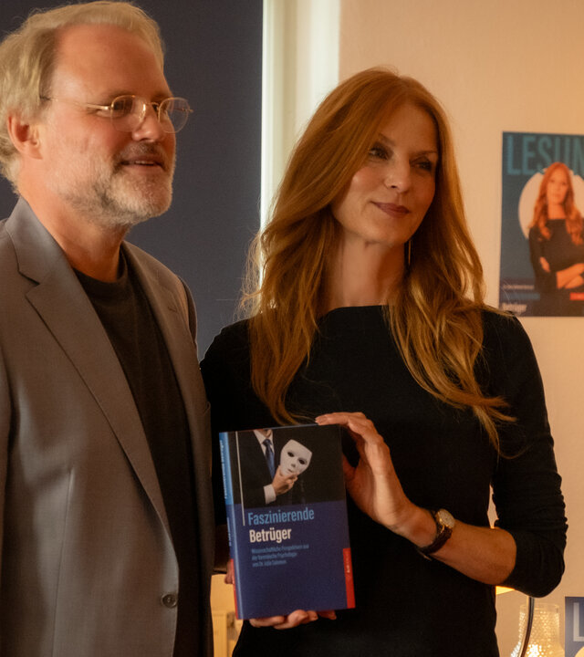 "Blutige Anfänger - Edelfeder": Henry Brill (Mirco Reseg) und Dr. Julia Salomon (Esther Schweins) stehen dicht nebeneinander in einem Raum. Julia hält ihr Buch vor ihrem Körper. Beide blicken lächelnd zur Seite.