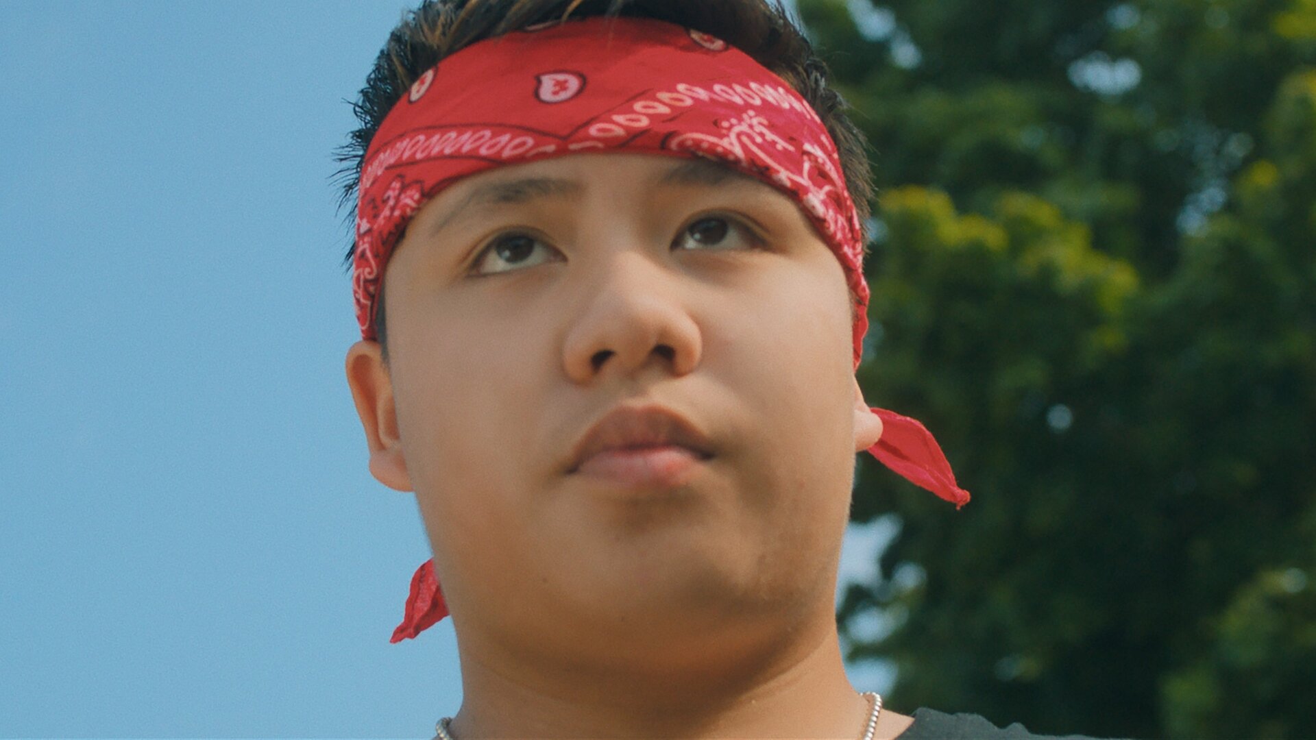 "Doppelhaushälfte - Roccos Rache": Untersicht: Rocco (Hoang Minh Ha) schaut mit ernstem Blick nach vorne und trägt ein rotes Bandana um den Kopf gebunden. Im Hintergrund steht eine Baumkrone vor blauem Himmel.