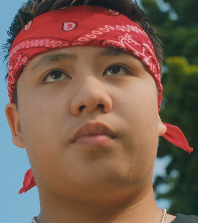 "Doppelhaushälfte - Roccos Rache": Untersicht: Rocco (Hoang Minh Ha) schaut mit ernstem Blick nach vorne und trägt ein rotes Bandana um den Kopf gebunden. Im Hintergrund steht eine Baumkrone vor blauem Himmel.