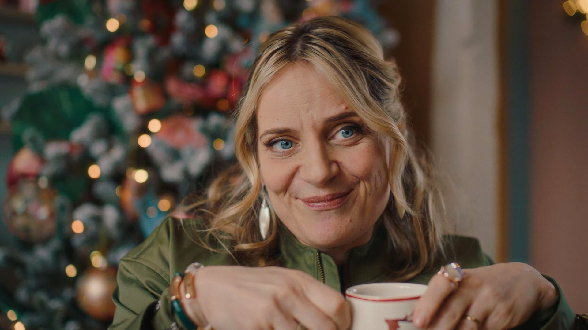 "Single Bells - Only Losers for Annie? - Rembert": Babs (Ruth Beeckmans) steht lächelnd vor einem üppig dekorierten Weihnachtsbaum. Sie hält eine Tasse mit beiden Händen fest.