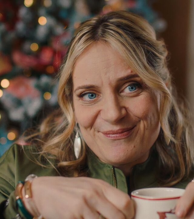 "Single Bells - Only Losers for Annie? - Rembert": Babs (Ruth Beeckmans) steht lächelnd vor einem üppig dekorierten Weihnachtsbaum. Sie hält eine Tasse mit beiden Händen fest.