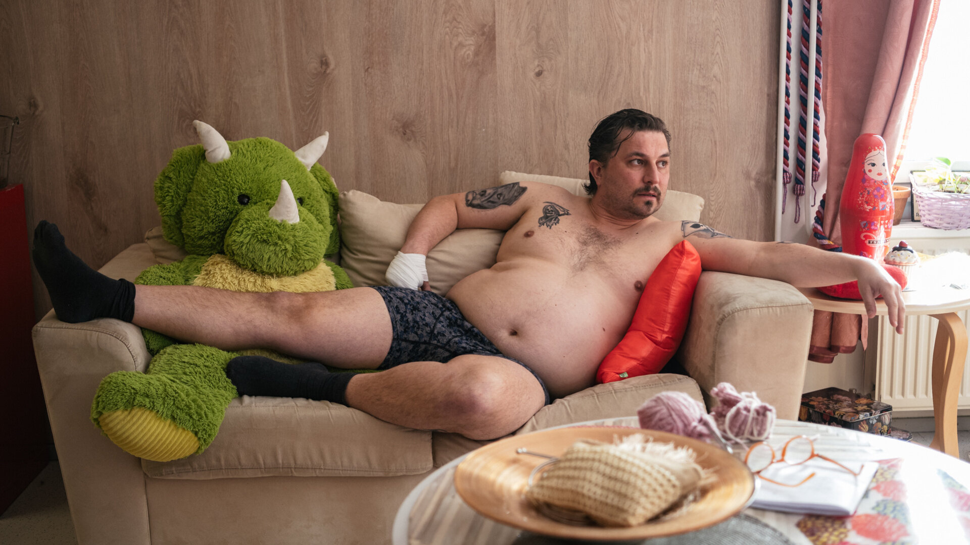 "Queen of Fucking Everything - Schmutzige Geschäfte": Spora (Joonas Heikkinen) liegt mit nackten Oberkörper auf einem Zweisitzer-Sofa. Er schaut zur Seite. Neben ihm sitzt ein großes Stofftier. An seiner rechten Hand trägt er einen Verband