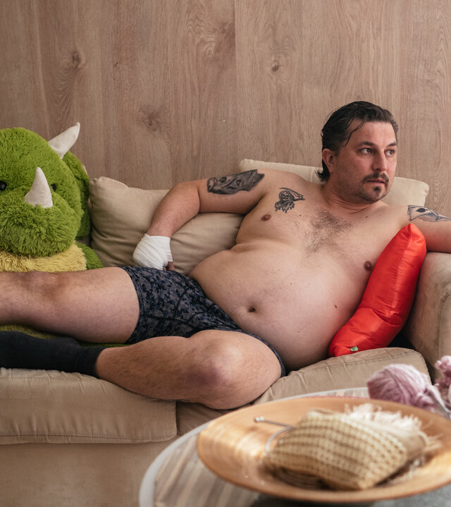 "Queen of Fucking Everything - Schmutzige Geschäfte": Spora (Joonas Heikkinen) liegt mit nackten Oberkörper auf einem Zweisitzer-Sofa. Er schaut zur Seite. Neben ihm sitzt ein großes Stofftier. An seiner rechten Hand trägt er einen Verband