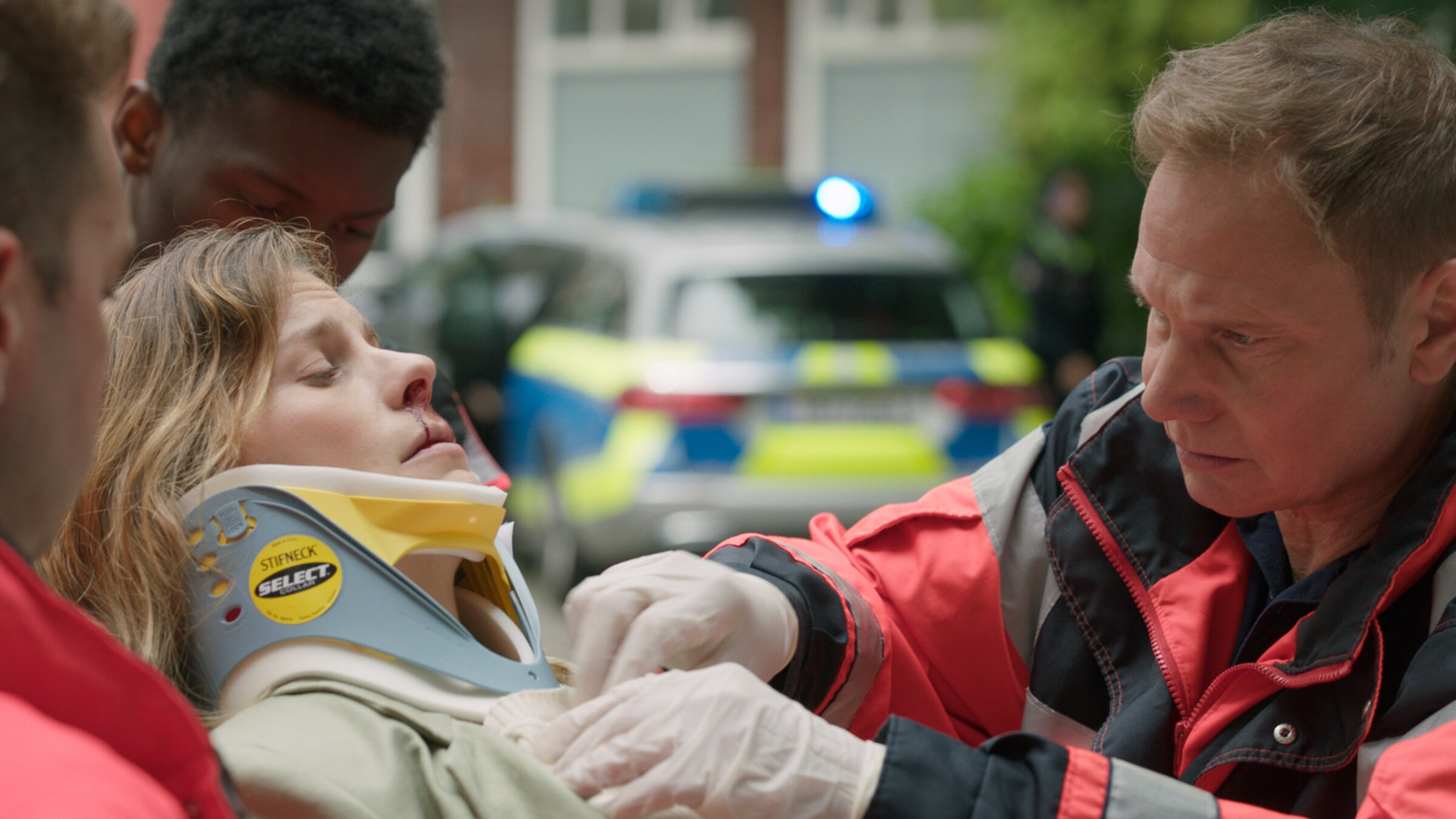 "Notruf Hafenkante - Kein Zurück": Großaufnahme von Dr. Haase (Fabian Harloff) und Julia Jacobi (Inga Lessmann). Dr. Haase versorgt Julia Jacobi, die auf einer Patiententrage liegt, daneben stehen zwei Sanitäter (Komparsen). Im Hintergrund steht ein Streifenwagen.