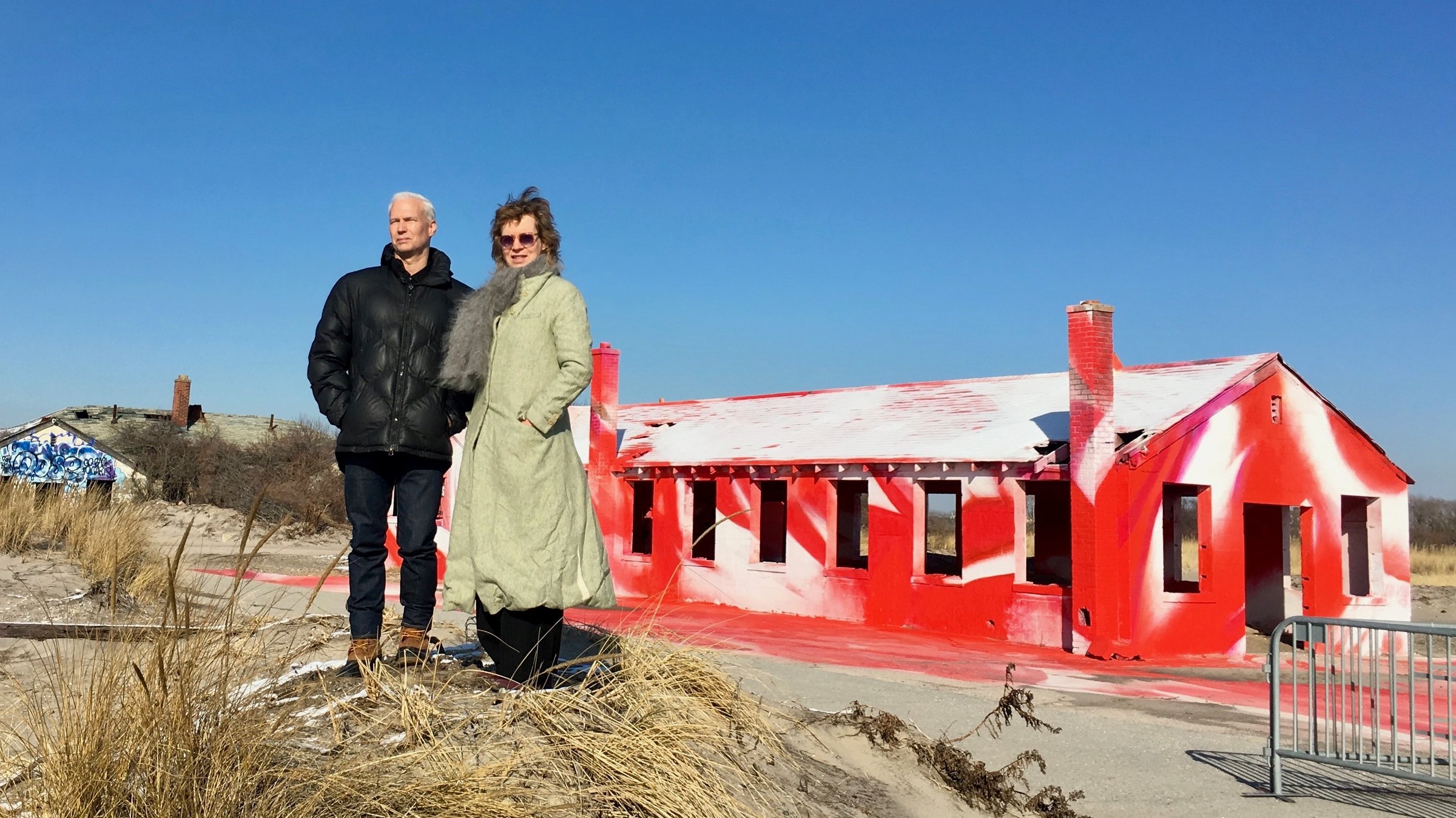 "Katharina Grosse - Think big!": Ein dunkelgekleideter Mann mit kurzen weißen Haaren und eine Frau mit hellem Mantel, Sonnenbrille und Schal stehen leicht erhöht auf einer grasbewachsenen Düne am Strand, im Hintergrund das Skelett eines verlassenen Hauses, das vollständig rot und weiß bemalt wurde.