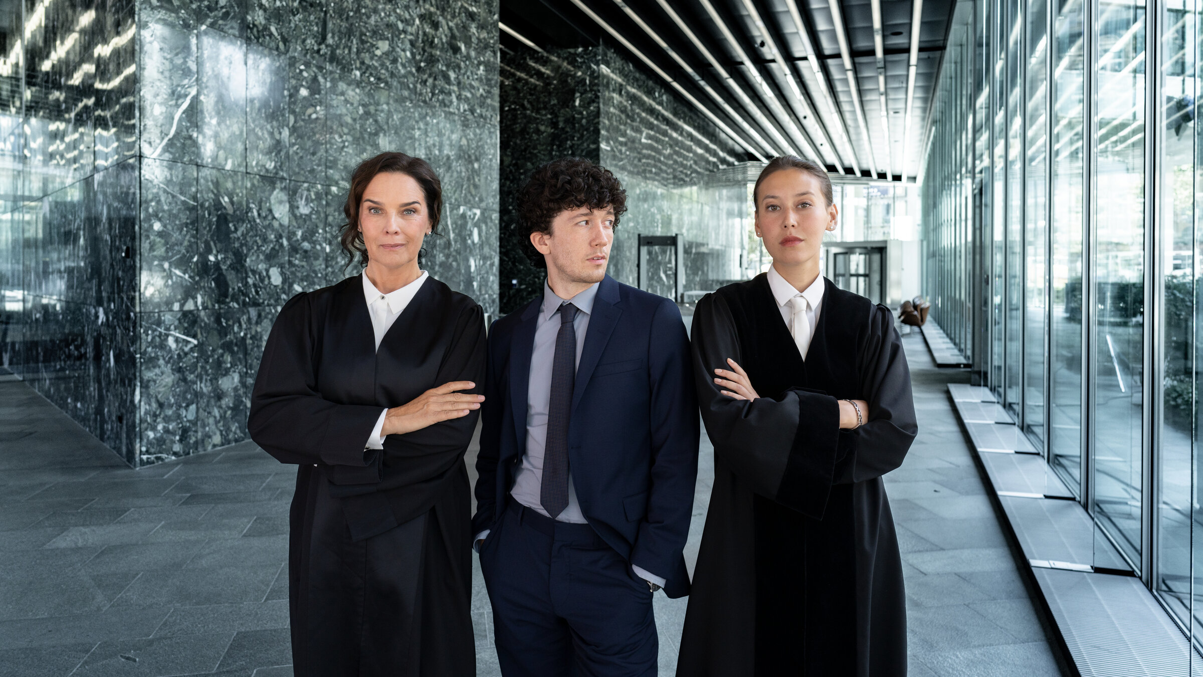 "Ein Fall für Conti - Spieler": Anna Conti (Désirée Nosbusch), Carlo von Hehenkamp (Maximilian Mundt) und Henry Mahn (Malaya Stern Takeda) stehen nebeneinander im großzügigen Foyer eines modernen Gebäudes.