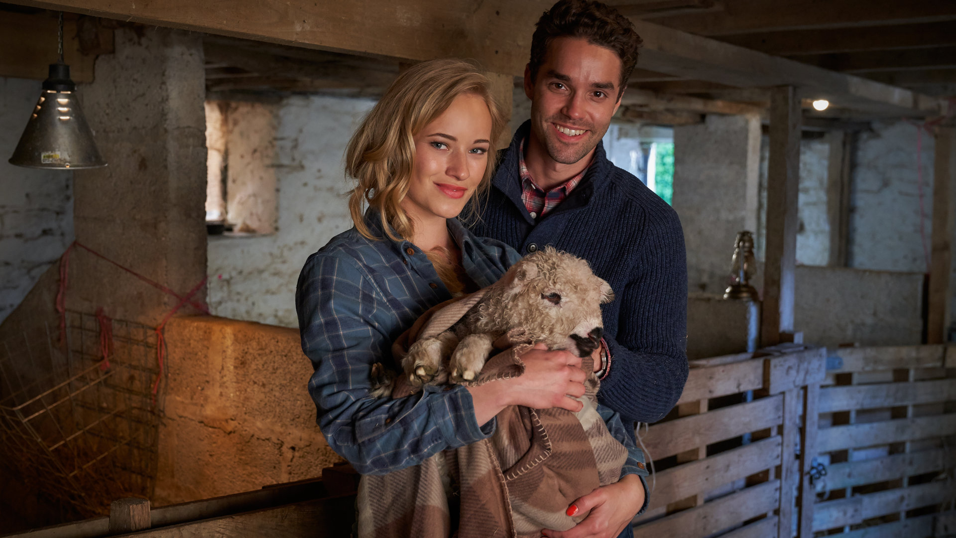 "Rosamunde Pilcher: Meine Cousine, die Liebe und ich": Holly (Valerie Huber) und Aaron (Max Befort) stehen gemeinsam im Stall und lächeln in die Kamera. Holly hält ein Lamm in eine Decke gewickelt auf dem Arm.
