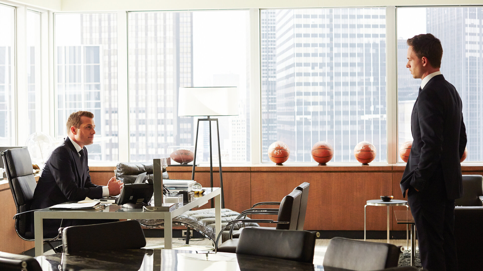 "Suits - Wissen, wann Schluss ist": Harvey (Gabriel Macht) sitzt an seinem Schreibtisch in einem hellen, modern eingerichteten Büro und blickt zu Mike (Patrick J. Adams), der mit ernster Miene im Raum steht. Im Hintergrund stehen mehrere Basketbälle auf einem Sideboard, dahinter ist die Skyline durch große Fenster zu sehen.