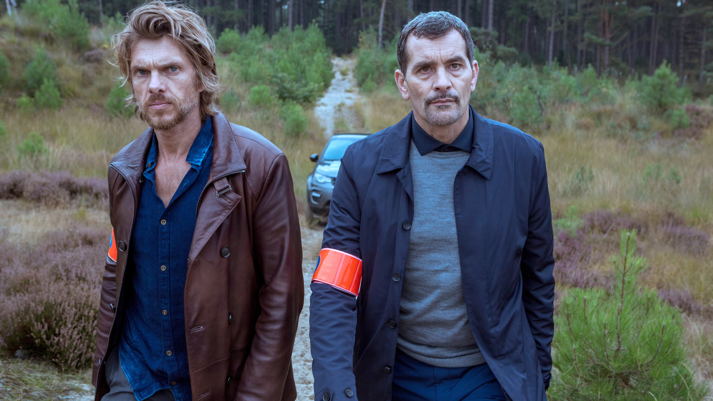 "Das letzte Opfer": Verstuyft (Werner De Smedt) und Vincke (Koen De Bouw) laufen über einen Feldweg auf die Kamera zu. Beide tragen eine rote Armbinde am rechten Arm. Im Hintergrund steht ihr Wagen auf einem Waldweg.