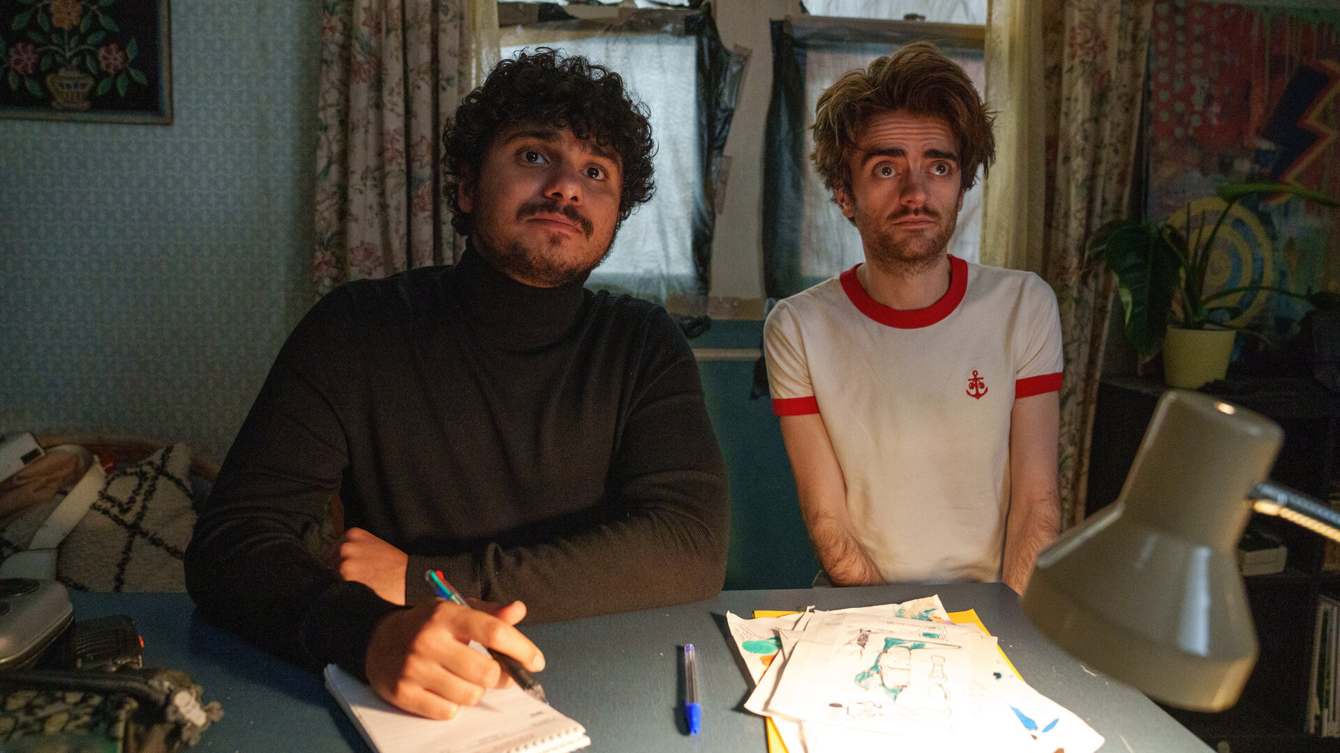 "Extraordinary - Sackgasse": Kash (Bilal Hasna) und Jizzlord (Luke Rollason) sitzen in einem Arbeitszimmer an einem Schreibtisch. Kash schaut aufmerksam geradeaus und hält dabei einen Stift in der Hand, der auf einem Block aufliegt, während Jizzlord skeptisch blickt.