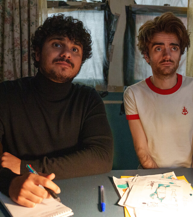 "Extraordinary - Sackgasse": Kash (Bilal Hasna) und Jizzlord (Luke Rollason) sitzen in einem Arbeitszimmer an einem Schreibtisch. Kash schaut aufmerksam geradeaus und hält dabei einen Stift in der Hand, der auf einem Block aufliegt, während Jizzlord skeptisch blickt.