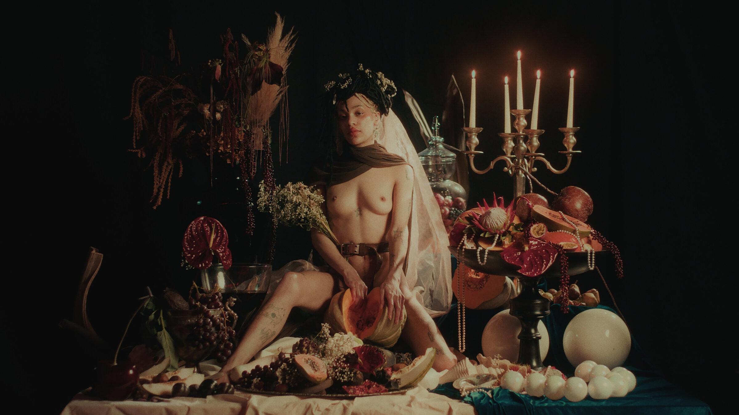 "Ab 18! - A Body Like Mine": Eine nackte junge Frau mit Blumenschmuck im Haar sitzt auf einem opulent arrangierten Tisch voller Früchte und Blumen.