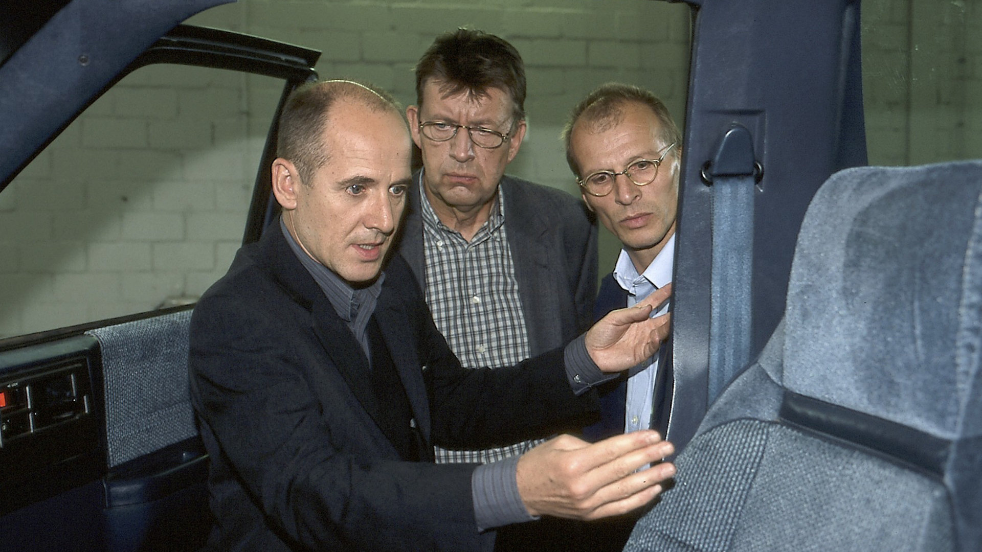 "Der letzte Zeuge - Im Netz": Dr. Robert Kolmaar (Ulrich Mühe), Joe Hoffer (Jörg Gudzuhn) und der Unfallsachverständiger Walter Erhardt (Jens-Uwe Bogadtke) stehen an einer Autotür und schauen alle drei ins Innere des Fahrzeugs.