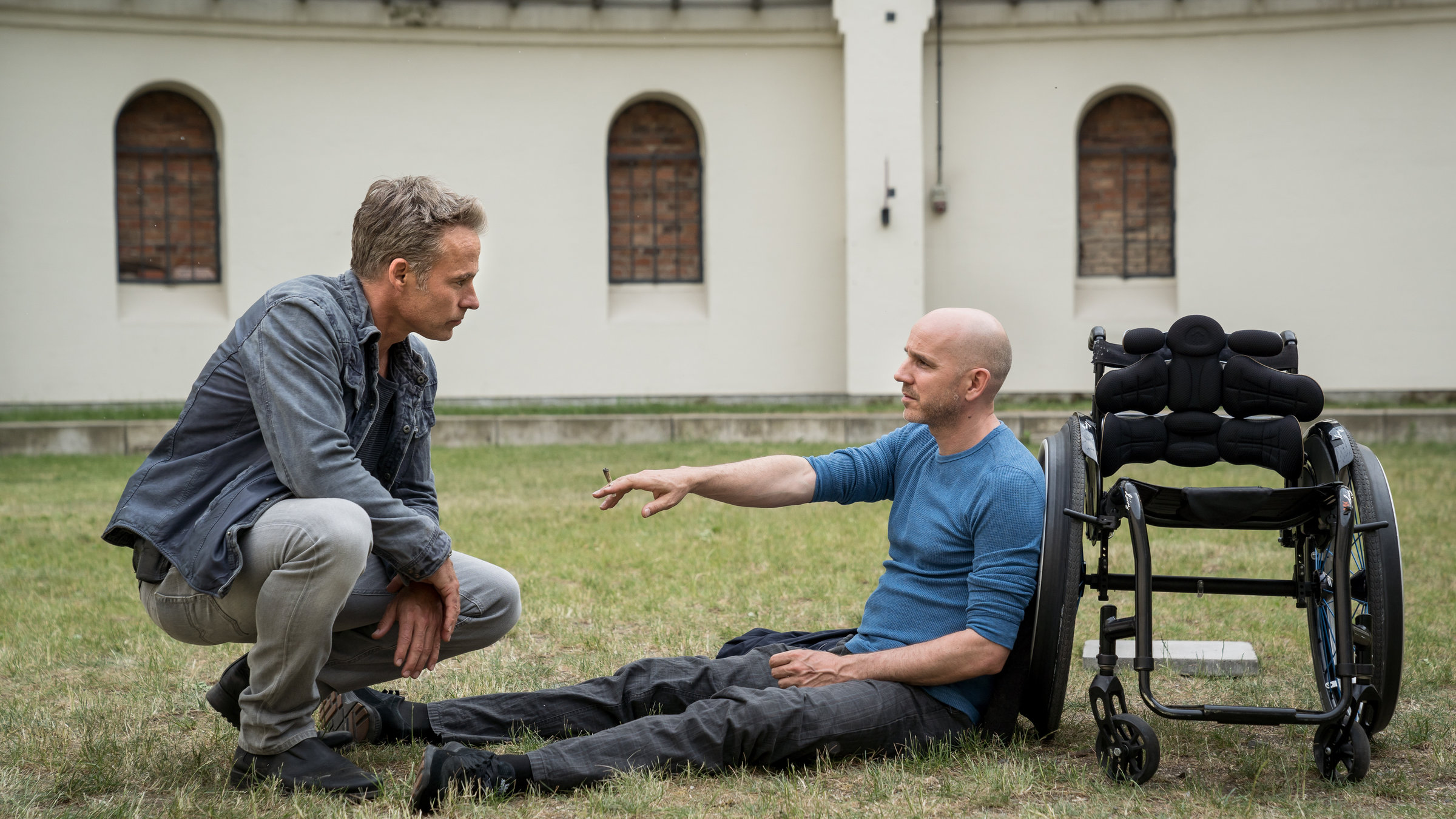 "SOKO Leipzig - Absturz": Jan Maybach (Marco Girnth) sitzt in Hocke gegenüber von Peter Hoffmeister (Paul Herwig), der auf einer Rasenfläche an seinen Rollstuhl gelehnt sitzt. Peter Hoffmeister streckt Jan Maybach seinen rechten Arm entgegen und hat einem Joint in der Hand. Im Hintergrund sieht man ein altes Industriegebäude.