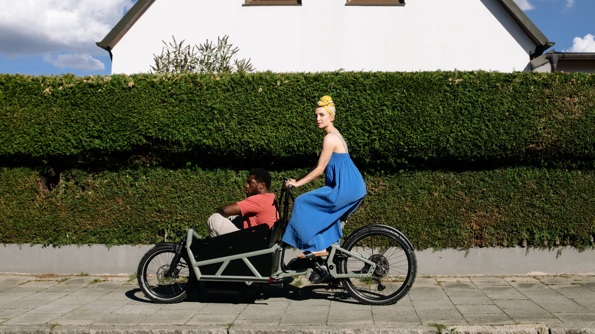 "I don't work here - Kitzbühel": Laura (Sina Martens) fährt in einem blauen Kleid auf einem schwarzen Lastenrad an einer grünen Hecke vorbei, hinter der ein weißes Haus mit zwei Fenstern zu sehen ist. Dawit (Akeem von Flodrop) sitzt mit angezogenen Beinen vorne im Lastenrad und lässt sich von Laura fahren, die zur Seite schaut.