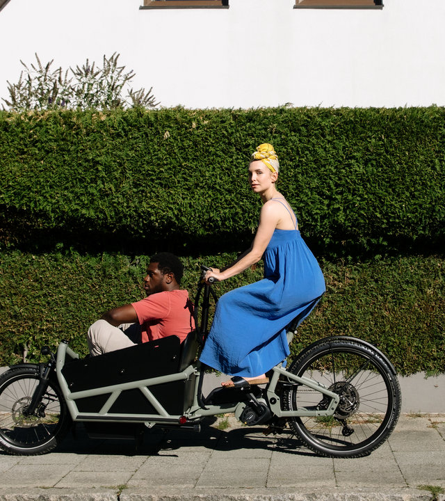 "I don't work here - Kitzbühel": Laura (Sina Martens) fährt in einem blauen Kleid auf einem schwarzen Lastenrad an einer grünen Hecke vorbei, hinter der ein weißes Haus mit zwei Fenstern zu sehen ist. Dawit (Akeem von Flodrop) sitzt mit angezogenen Beinen vorne im Lastenrad und lässt sich von Laura fahren, die zur Seite schaut.