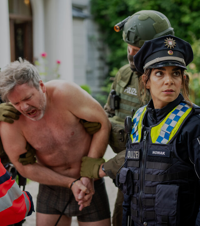"Notruf Hafenkante - Gefährliche Lage": Zwei SEK Beamte (Komparsen) halten Lars Markwardt (Nicholas Reinke) in ihrer Mitte fest. Markwardt trägt nur eine Unterhose. Sein Gesicht ist schmerzverzerrt, seine Hände sind mit einem Kabelbinder zusammengebunden. Vor ihm steht Dr. Jordan (Hanife Sylejmani). Seitlich neben ihr steht die Polizistin Isa Nowak (Lilli Hollunder) und ihr Kollege Kris Freiberg (Marc Barthel).