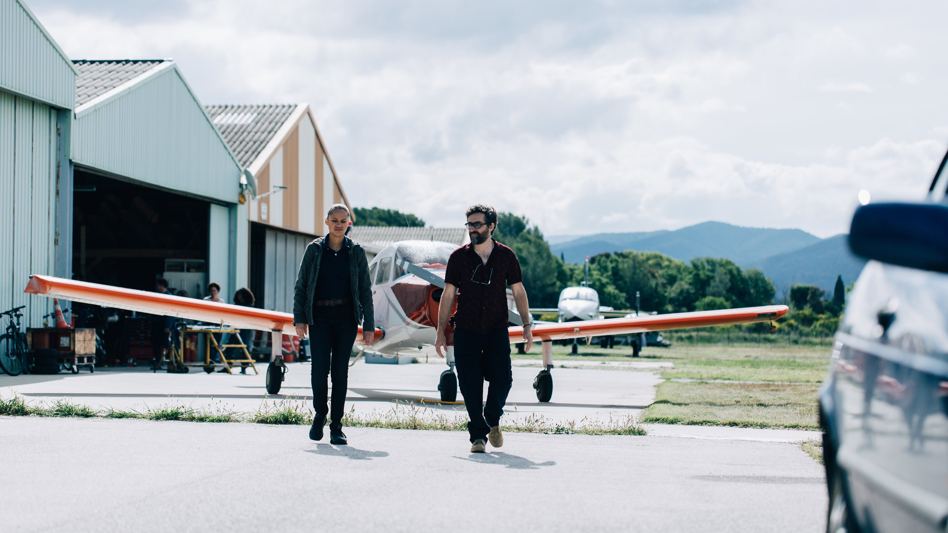 "They Were Ten": Außenaufnahme eines Flugzeughangars mit zwei Flugzeugen davor. Vor dem vorderen Flugzeug gehen Bapista (Wendy Nieto) und Mathieu (Mathieu Demy) in Richtung Kamera auf ein Auto zu, das am rechten Bildrand unscharf angeschnitten ist.
