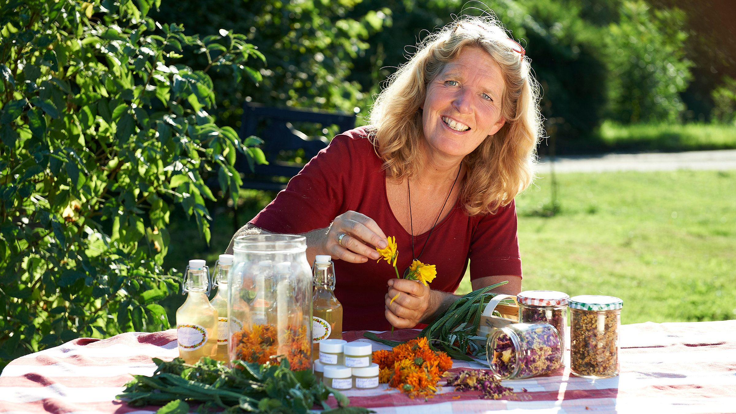 "Lecker aufs Land - Eine kulinarische Herbstreise - Die Wildkräuter-Lady aus dem Bergischen Land": Petra Burgmer sitzt am Tisch im Freien, vor ihr liegen Kräuter.