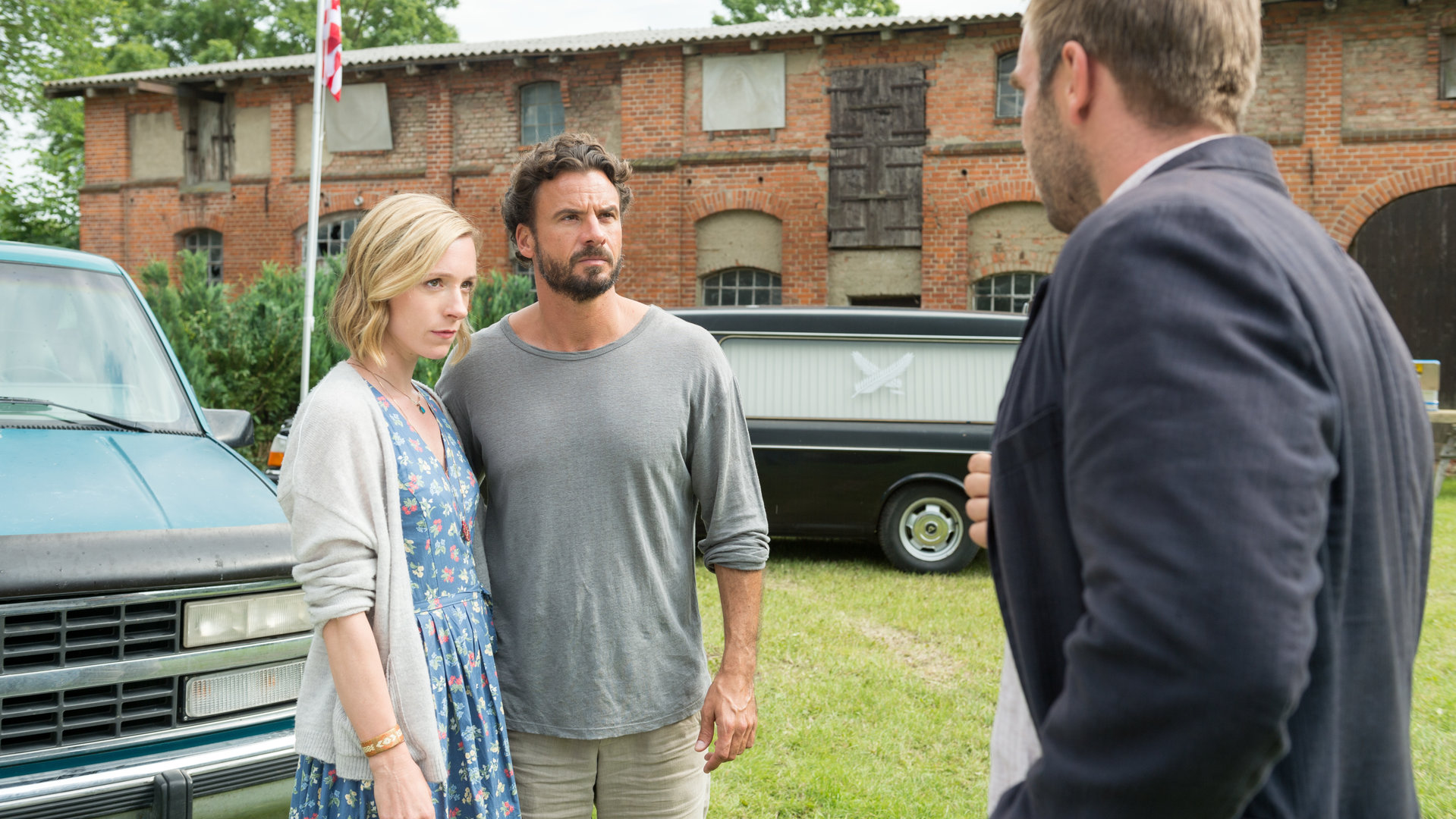"SOKO Wismar – Ein nachhaltiger Tod": Julia Fedder (Friederike Linke) und Mark Fedder (Stephan Luca) und  stehen Arm in Arm auf dem Hof. Vor ihnen steht Kommissar Lars Pöhlmann (Dominic Boeer). Im Hintergrund sieht man einen schwarzen Leichenwagen vor einer alten Scheune stehen.