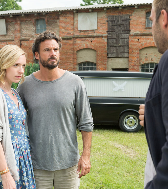 "SOKO Wismar – Ein nachhaltiger Tod": Julia Fedder (Friederike Linke) und Mark Fedder (Stephan Luca) und  stehen Arm in Arm auf dem Hof. Vor ihnen steht Kommissar Lars Pöhlmann (Dominic Boeer). Im Hintergrund sieht man einen schwarzen Leichenwagen vor einer alten Scheune stehen.