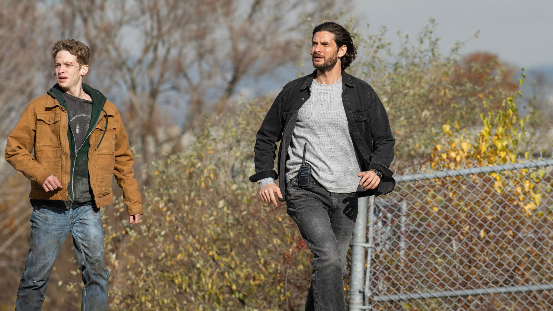 "Das Institut - Die Hölle wartet": Luke (Joe Freeman) und Tim (Ben Barnes) sind im Freien unterwegs. Sie wirken gehetzt. Im Hintergrund blüht ein Busch hinter einem Maschendrahtzaun.