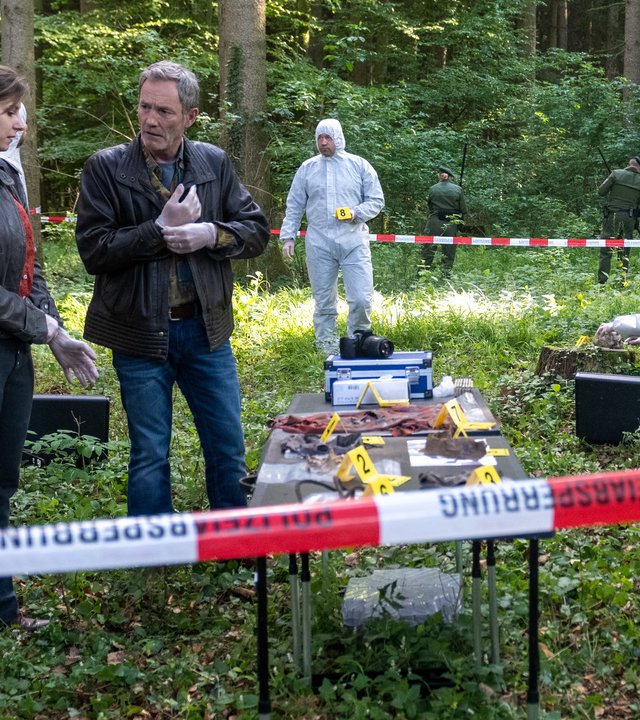 "XY gelöst": Nachgespielte Szene in einem Waldstück dass von der Polizei abgesperrt ist. Mitten drin steht ein Tisch mit Fundsachen. Sie sind nummeriert. Eine Frau und ein Mann ziehen sich Gummihandschuhe an und beraten sich. Zwei Kollegen der Spurensicherung in weißen Overalls stehen dabei.