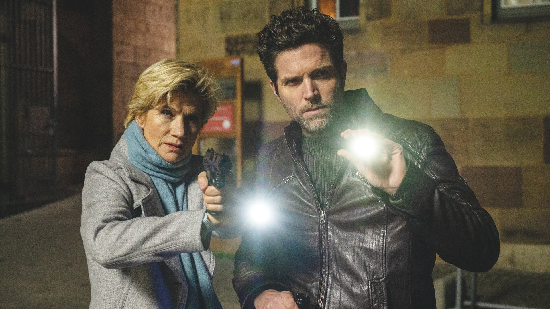 "SOKO Stuttgart - Dunkelheit (1)": Erste Kriminalhauptkommissarin Martina Seiffert (Astrid M. Fünderich) und Kriminalhauptkommissar Jo Stoll (Peter Ketnath) stehen im Dunkeln vor einem Gebäude. Sie halten Taschenlampen in den Händen und blicken beide gezielt in eine Richtung. Martina hat zudem ihre Dienstwaffe gezogen.