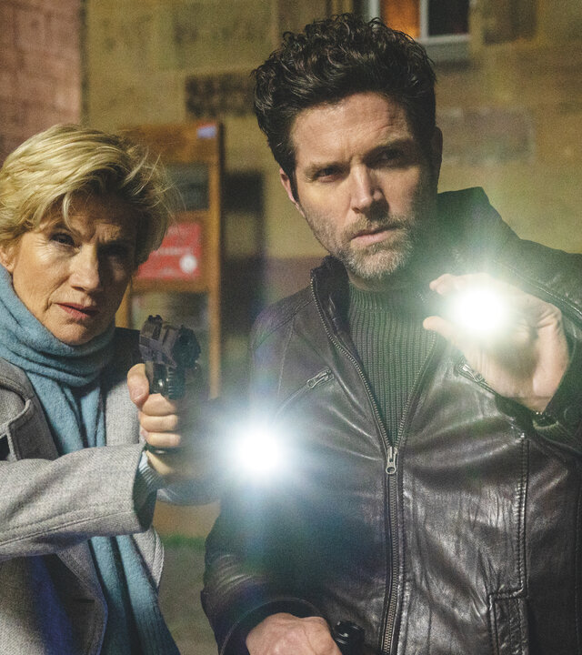 "SOKO Stuttgart - Dunkelheit (1)": Erste Kriminalhauptkommissarin Martina Seiffert (Astrid M. Fünderich) und Kriminalhauptkommissar Jo Stoll (Peter Ketnath) stehen im Dunkeln vor einem Gebäude. Sie halten Taschenlampen in den Händen und blicken beide gezielt in eine Richtung. Martina hat zudem ihre Dienstwaffe gezogen.