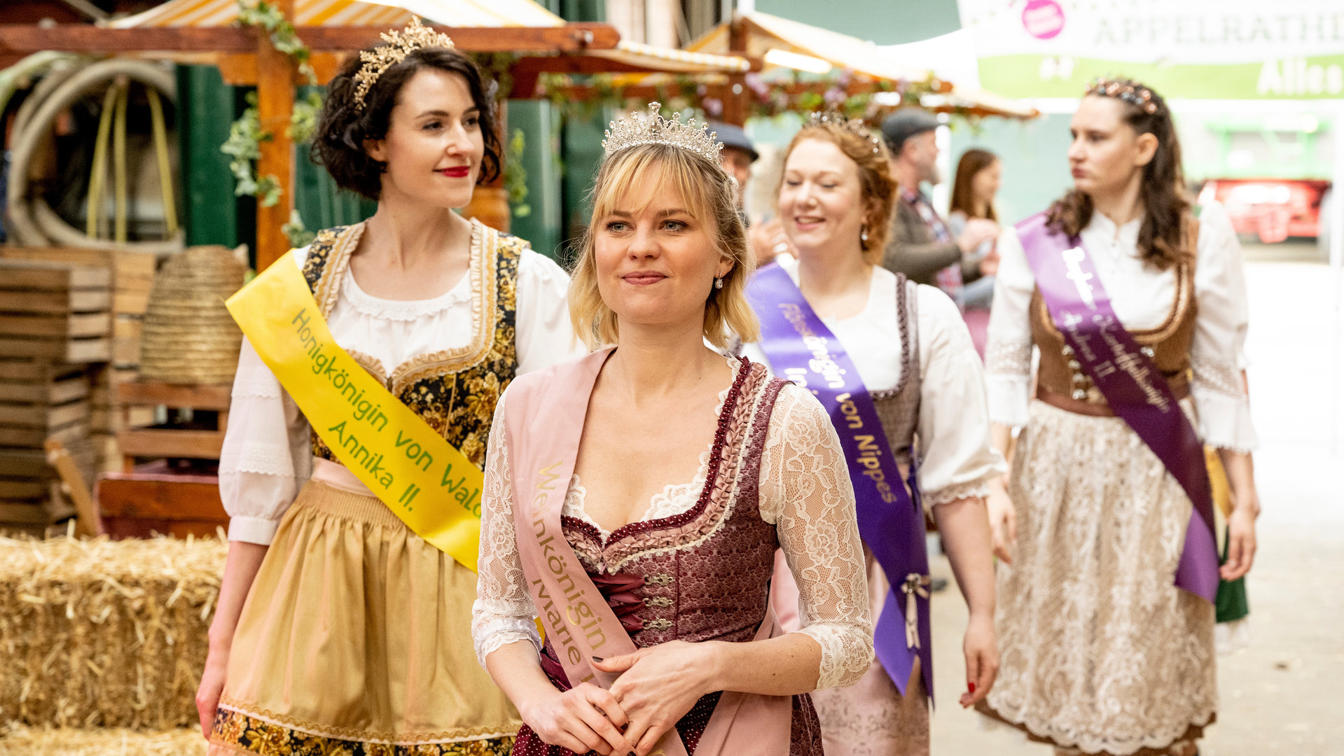 "SOKO Köln - Die Milchkönigin von Appelrath": Marie Neffgen (Henrike Fehrs) läuft in einem, mit Spitzen verzierten, weinroten Dirndl lächelnd über einen Hof. Sie trägt eine rosafarbene Scherpe quer über dem Körper, auf der unter anderem die Worte "Weinkönigin" und "Marie" zu lesen sind. Außerdem sitzt auf ihrem Kopf eine Krone. Hinter ihr laufen drei weitere Frauen (Komparsinnen) in Dirndln, die ebenfalls Scherpen und Kronen tragen. Im Hintergrund stehen drei weitere Personen (Komparsen).