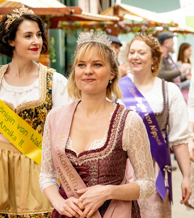 "SOKO Köln - Die Milchkönigin von Appelrath": Marie Neffgen (Henrike Fehrs) läuft in einem, mit Spitzen verzierten, weinroten Dirndl lächelnd über einen Hof. Sie trägt eine rosafarbene Scherpe quer über dem Körper, auf der unter anderem die Worte "Weinkönigin" und "Marie" zu lesen sind. Außerdem sitzt auf ihrem Kopf eine Krone. Hinter ihr laufen drei weitere Frauen (Komparsinnen) in Dirndln, die ebenfalls Scherpen und Kronen tragen. Im Hintergrund stehen drei weitere Personen (Komparsen).