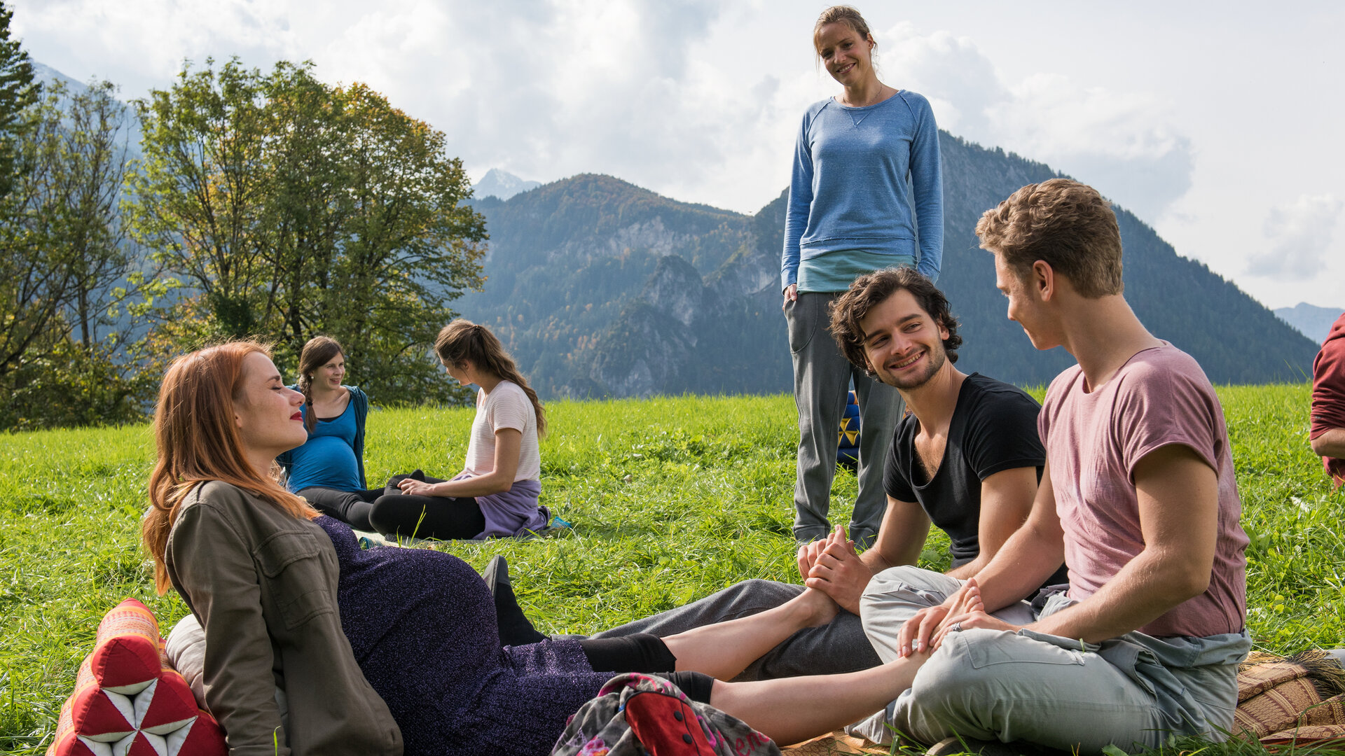 "Lena Lorenz - Zwei Väter (1)": Auf einer grünen Wiese vor einem Bergpanorama sitzen Anna Kraus (Anna Hausburg), Paul Mannhardt (Aaron Karl) und Max König (Rafael Gareisen). Die beiden Männer massieren Annas Füße, Lena Lorenz (Patricia Aulitzky) schaut ihnen stehend  zu. Anna genießt die Fußmassage. Im Hintergrund sitzen zwei Frauen (Komparsinnen).
