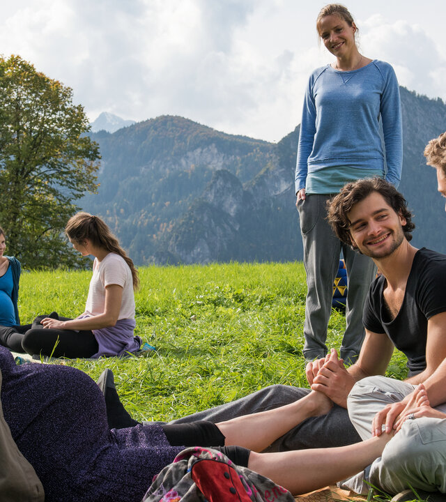 "Lena Lorenz - Zwei Väter (1)": Auf einer grünen Wiese vor einem Bergpanorama sitzen Anna Kraus (Anna Hausburg), Paul Mannhardt (Aaron Karl) und Max König (Rafael Gareisen). Die beiden Männer massieren Annas Füße, Lena Lorenz (Patricia Aulitzky) schaut ihnen stehend  zu. Anna genießt die Fußmassage. Im Hintergrund sitzen zwei Frauen (Komparsinnen).