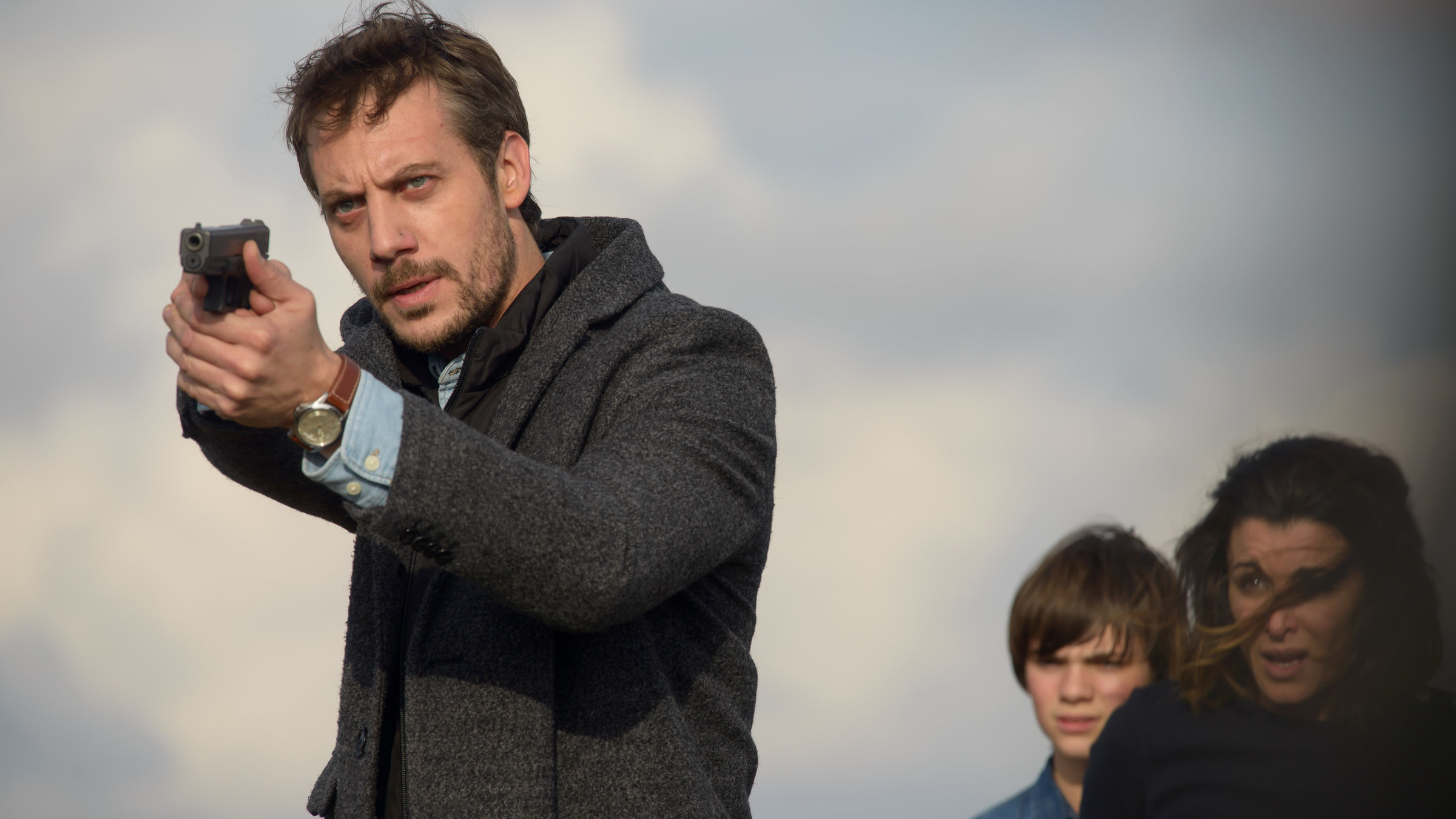 "Zwei auf der Flucht": Donatelli (Joffrey Platel) steht mit gezogener Schusswaffe im Freien und zielt ins Off. Hinter ihm sind Sarah (Jenifer Bartoli) und Léo (Félix Bossuet) zu sehen, die einen ängstlichen Gesichtsausdruck haben.