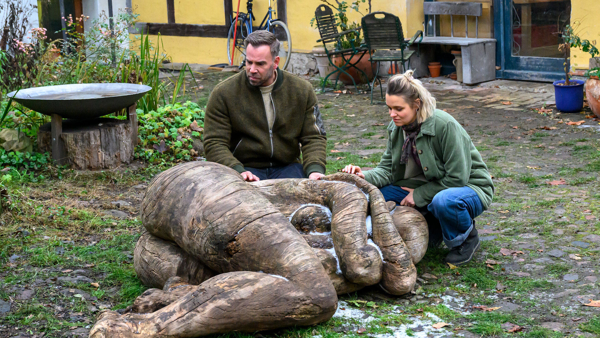 „SOKO Wismar – Königin der Hölzer“: Lars Pöhlmann (Dominic Boeer) betrachtet im Hof der Bildhauer fasziniert eine Holzskulptur, die eine menschliche Figur in Embryonalstellung zeigt. Neben ihm kniet Lilly Kladow (Nora Schulte) und legt die Hand auf die Schulter der Skulptur. Im Hintergrund befindet sich die Bildhauerwerkstatt.