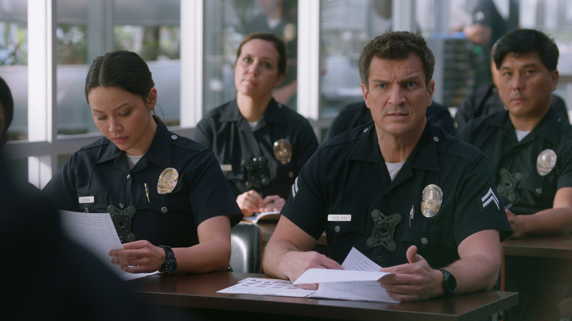 "The Rookie - Armee der Freiheit": Officer Lucy Chen (Melissa O'Neil) und Officer Nolan (Nathan Fillion) sitzen an einem Tisch in einem Besprechungsraum und halten jeweils Unterlagen in ihren Händen.