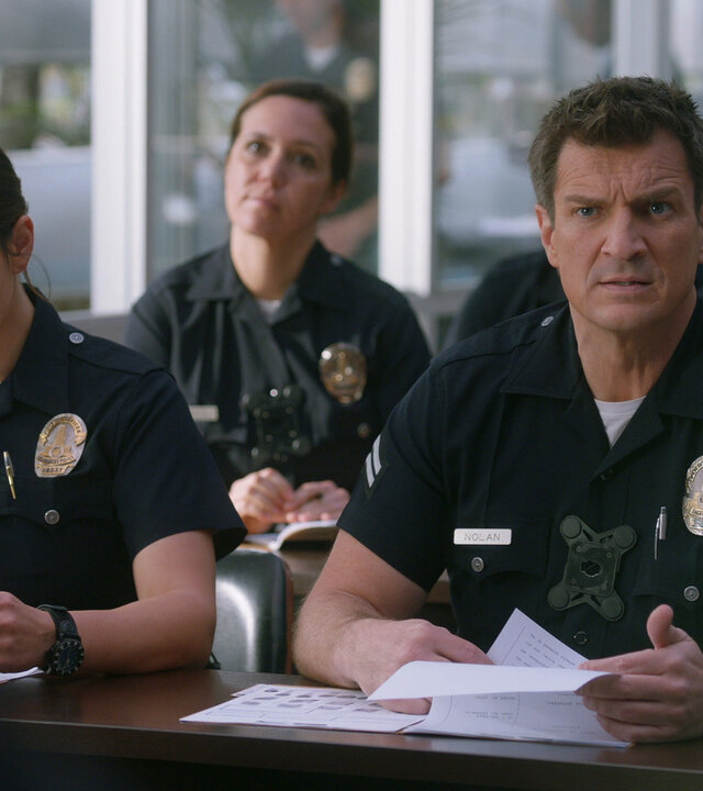 "The Rookie - Armee der Freiheit": Officer Lucy Chen (Melissa O'Neil) und Officer Nolan (Nathan Fillion) sitzen an einem Tisch in einem Besprechungsraum und halten jeweils Unterlagen in ihren Händen.