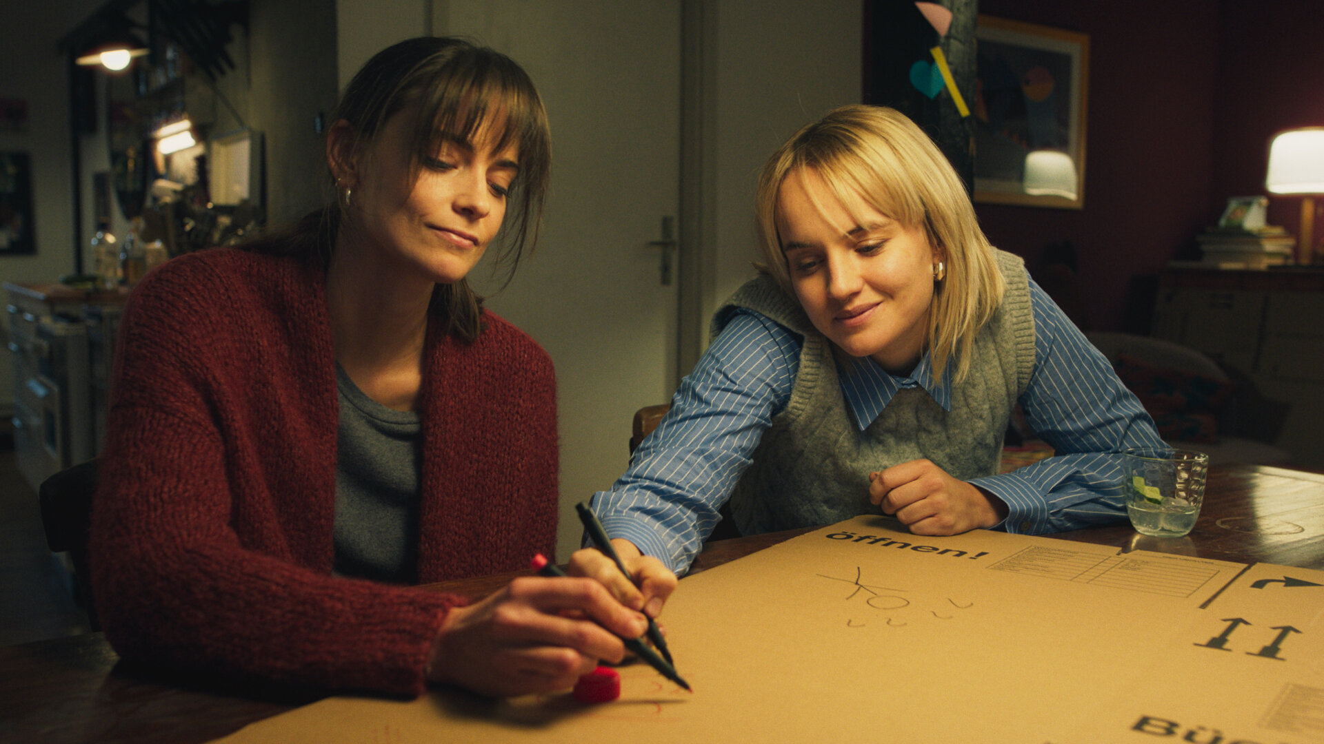 "Bauchgefühl - Tina": Lena (Laura Berlin) und Tina (Luise von Finckh) sitzen an einem Tisch und zeichnen mit Filzstiften Strichmännchen auf einen gefalteten Umzugskarton.