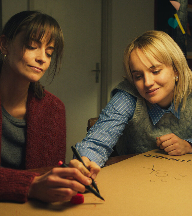"Bauchgefühl - Tina": Lena (Laura Berlin) und Tina (Luise von Finckh) sitzen an einem Tisch und zeichnen mit Filzstiften Strichmännchen auf einen gefalteten Umzugskarton.