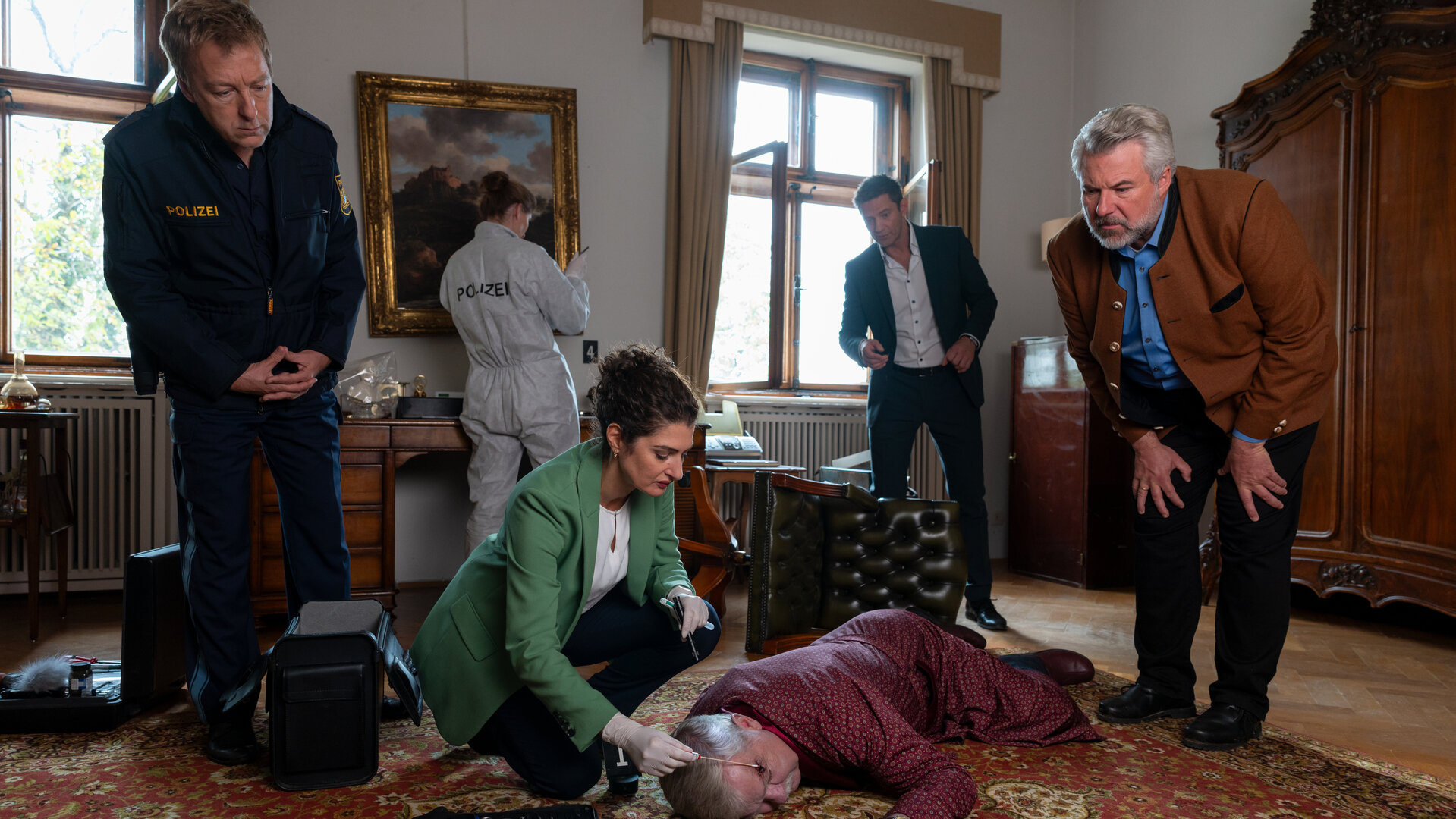"Die Rosenheim-Cops - Spuk in Rosenheim": Dr. Dimos (Anastasia Papadopoulou) kniet auf einem rotgemusterten Perserteppich und untersucht das Opfer Gustav von Hausenring (Komparse), das bäuchlings vor ihr liegt. Um sie herum stehen Polizeihauptmeister Mohr (Max Müller), Kommissar Hansen (Igor Jeftić) und Kommissar Stadler (Dieter Fischer), die sich vorbeugen, um das Opfer zu betrachten. Im Hintergrund arbeitet eine Kollegin (Komparsin) der Polizei in weißem Schutzanzug, die mit dem Rücken zur Kamera gewandt vor einem Gemälde steht, das an der Wand hängt.