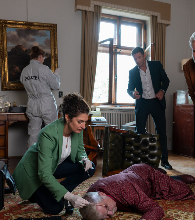 "Die Rosenheim-Cops - Spuk in Rosenheim": Dr. Dimos (Anastasia Papadopoulou) kniet auf einem rotgemusterten Perserteppich und untersucht das Opfer Gustav von Hausenring (Komparse), das bäuchlings vor ihr liegt. Um sie herum stehen Polizeihauptmeister Mohr (Max Müller), Kommissar Hansen (Igor Jeftić) und Kommissar Stadler (Dieter Fischer), die sich vorbeugen, um das Opfer zu betrachten. Im Hintergrund arbeitet eine Kollegin (Komparsin) der Polizei in weißem Schutzanzug, die mit dem Rücken zur Kamera gewandt vor einem Gemälde steht, das an der Wand hängt.