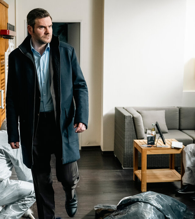 "SOKO Köln - Wer Kollegen hat, braucht keine Feinde": Jonas Fischer (Lukas Piloty) läuft in einem Wohnraum neben einer Leiche, die mit einer dunkelgrauen Plane abgedeckt wurde. Er sieht verwundert zur Seite. Im Hintergrund sitzt auf einem hellgrauen Sofa Dr. Pilar Westphal (Yulia Yáñez Schmidt), die vor sich auf ein Tablet sieht. Hinter Jonas kniet ein Mitarbeiter (Komparse) der Spurensicherung. Neben diesem steht ein aufgeklappter Koffer mit Arbeitsmaterialien. An der Seite steht ein weiterer Mitarbeiter (Komparse) der Spurensicherung.