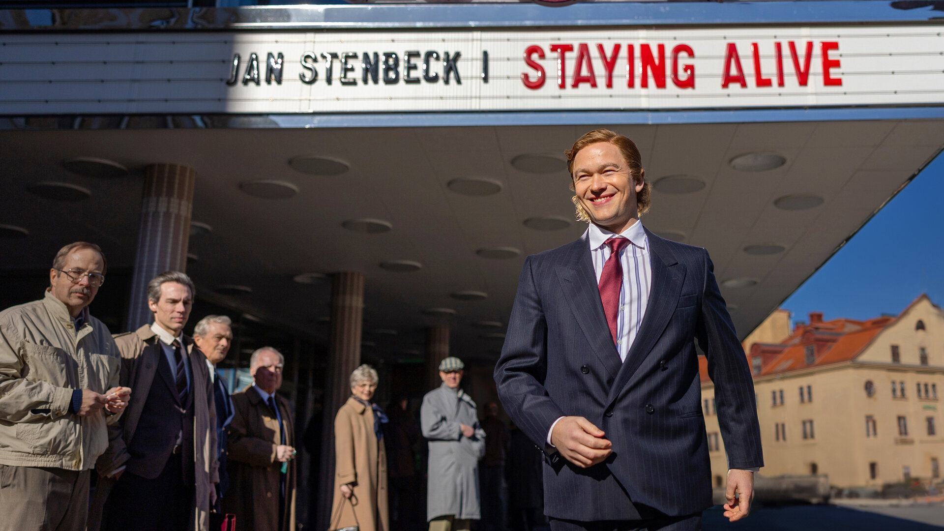 "Stenbeck - Staying Alive - Kampf um Kontrolle": Ein lachender Jan Stenbeck (Jabob Oftebro) kommt unter dem überdachten Eingangsbereich eines Kinos hervorgelaufen, Passanten (Komparsen) schauen sich zu ihm um.