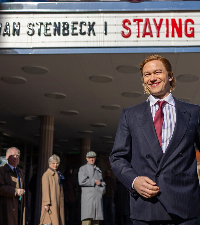 "Stenbeck - Staying Alive - Kampf um Kontrolle": Ein lachender Jan Stenbeck (Jabob Oftebro) kommt unter dem überdachten Eingangsbereich eines Kinos hervorgelaufen, Passanten (Komparsen) schauen sich zu ihm um.
