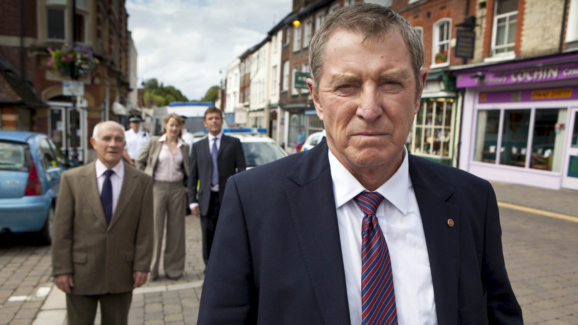 "Inspector Barnaby - Köpfen ist auch keine Lösung": Inspector Barnaby (John Nettles) steht auf einer Straße und schaut ernst. Im Hintergrund beobachten ihn Dr. George Bullard (Barry Jackson), DC Gail Stephens (Kirsty Dillon) und DS Ben Jones (Jason Hughes).