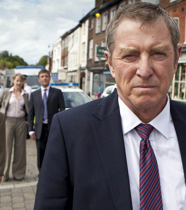 "Inspector Barnaby - Köpfen ist auch keine Lösung": Inspector Barnaby (John Nettles) steht auf einer Straße und schaut ernst. Im Hintergrund beobachten ihn Dr. George Bullard (Barry Jackson), DC Gail Stephens (Kirsty Dillon) und DS Ben Jones (Jason Hughes).