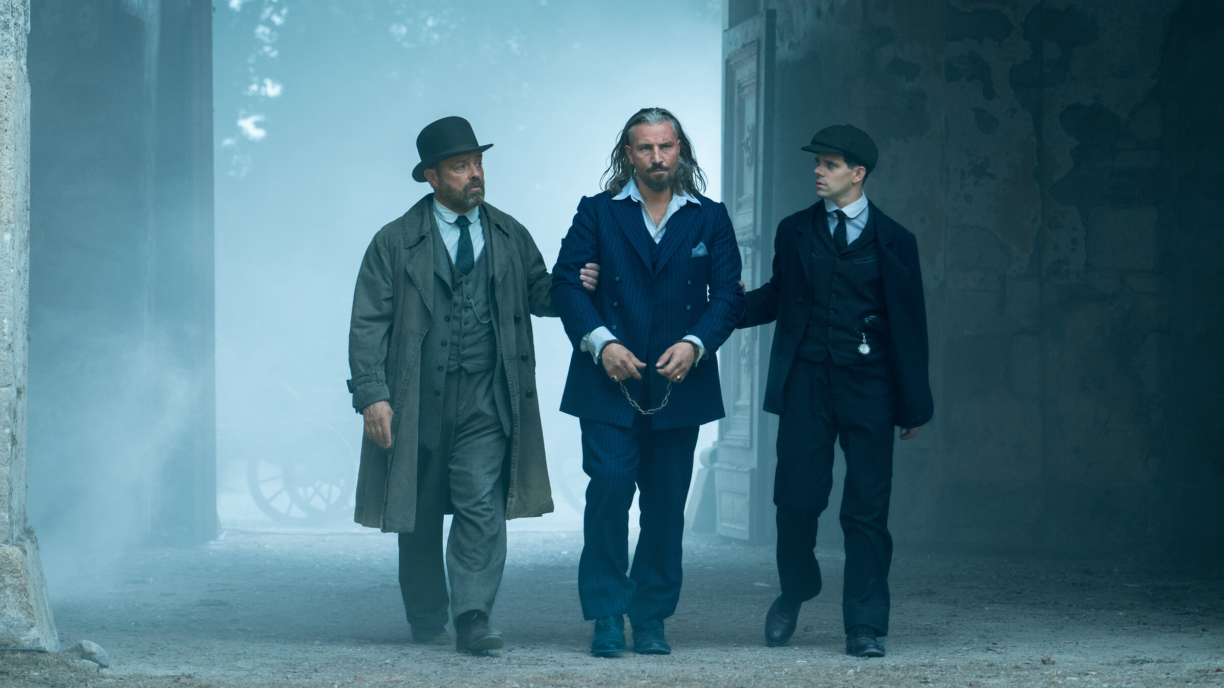 "Vienna Blood - Mephisto Teil 1": Oskar Rheinhardt (Juergen Maurer) und Haussmann (Josef Ellers) führen Franz Burgstaller (Murathan Muslu) in Handschellen durch ein Tor, dahinter schwebt Nebel.