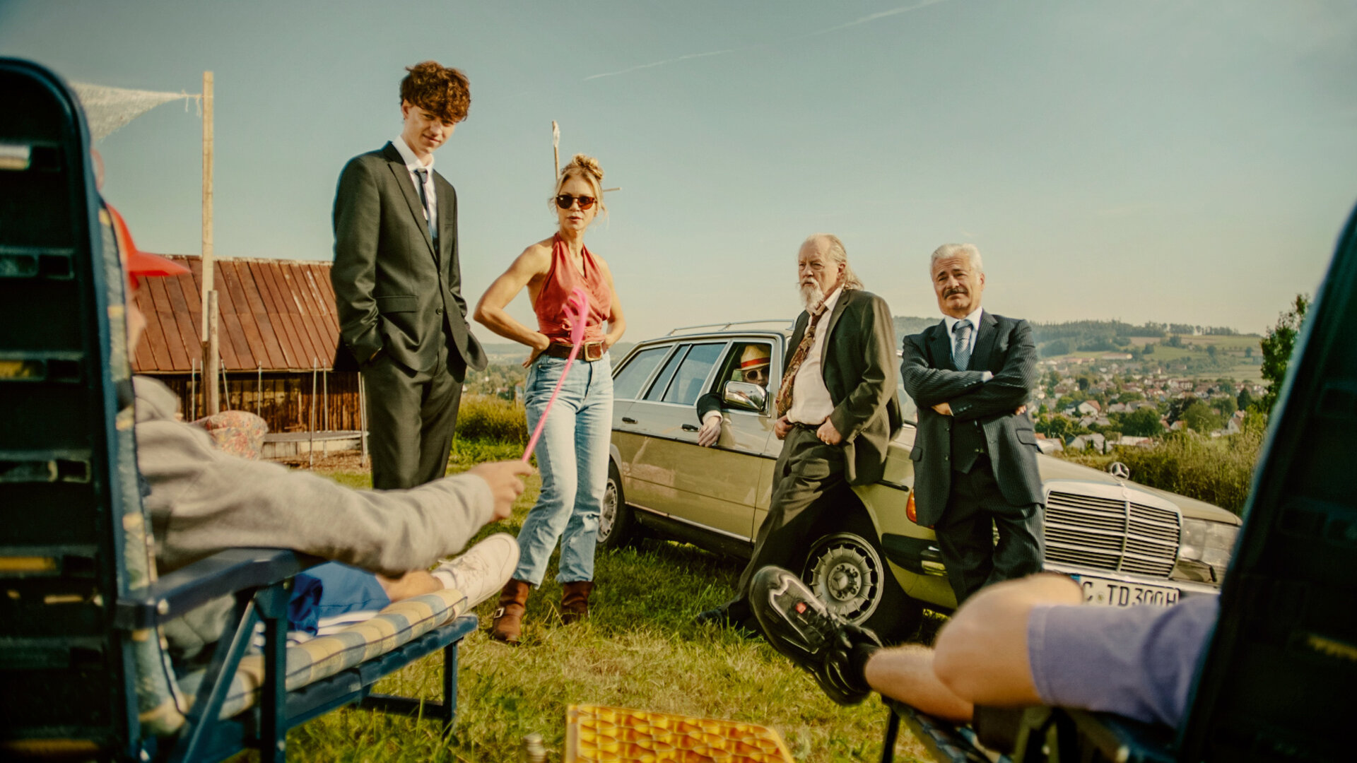 "Tschappel - Am Tag, als Carlo Brenner starb": Vor einem alten Bauwagen stehen Carlo (Jeremias Meyer) im dunklen Anzug und Gabi (Nina Gnädig) nebeneinander. Neben ihnen lehnen Hartmut (Paul Faßnacht) und Roberto (Martin Umbach) mit verschränkten Armen an ihrem alten Mercedes-Kombi, während Konrad (Gerhard Georg Jilka) mit Sonnenhut und Sonnenbrille aus dem Fenster des Wagens blickt. An den Seiten im Anschnitt stehen Liegestühle auf denen Personen sitzen die nicht zu erkennen sind.