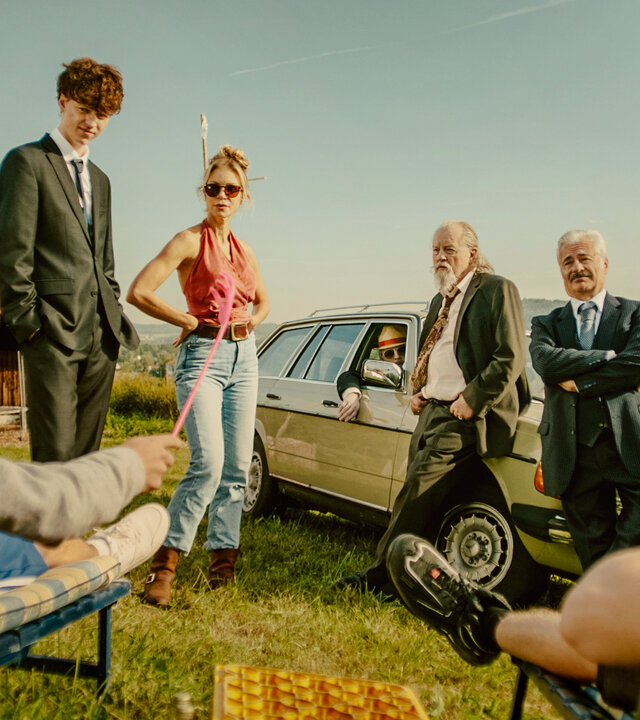 "Tschappel - Am Tag, als Carlo Brenner starb": Vor einem alten Bauwagen stehen Carlo (Jeremias Meyer) im dunklen Anzug und Gabi (Nina Gnädig) nebeneinander. Neben ihnen lehnen Hartmut (Paul Faßnacht) und Roberto (Martin Umbach) mit verschränkten Armen an ihrem alten Mercedes-Kombi, während Konrad (Gerhard Georg Jilka) mit Sonnenhut und Sonnenbrille aus dem Fenster des Wagens blickt. An den Seiten im Anschnitt stehen Liegestühle auf denen Personen sitzen die nicht zu erkennen sind.